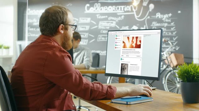 Creative Office Worker Browsing Social Network Pages, He Scrolls Through His Wall on a Personal Computer. In the Background Stylish Creative Agency Room. Shot on RED EPIC-W 8K Helium Cinema Camera.