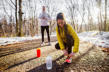 Close up of sporty active beautiful smiling slim woman in sportswear kneeling on the road and tying shoelaces in the sunny winter morning outside in nature with a handsome trainer behind her.