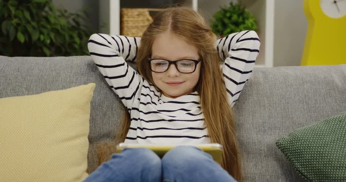 Portrait shot of the nice little girl in the glasses sitting on the sofa and watching something on her tablet device in the cozy room. Inside