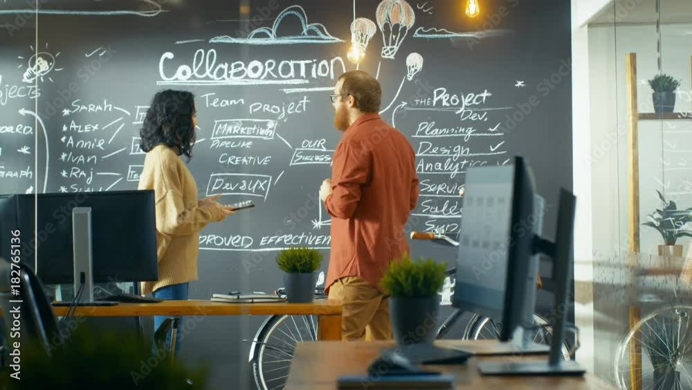 Poster female project manager holds tablet computer and shows plan drawn on a blackboard wall to male devel