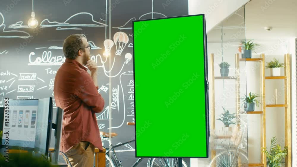 Poster male developer looks at interactive whiteboard with mock-up green screen. he smooths his beard think