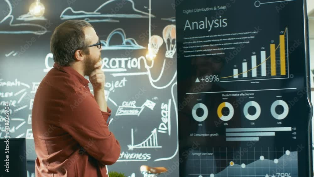 Poster Marketer Looks at Interactive Touchscreen Whiteboard Showing Latest Graphs and Charts on Statistical Growth. He Smooths His Beard in Thinking Gesture. Shot on RED EPIC-W 8K Helium Cinema Camera.