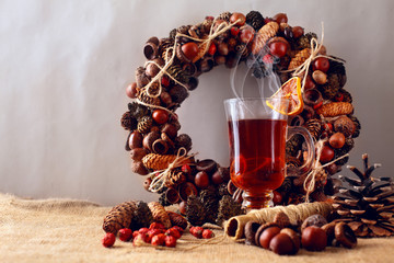 A glass of a hot spicy tea drink with dried lemon.Christmas wreath of arcorns and cones on second...