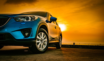 Blue compact SUV car with sport and modern design parked on concrete road by sea beach at sunset. New shiny SUV car drive for travel on summer vacations with road trip. Front view of electric car. - obrazy, fototapety, plakaty