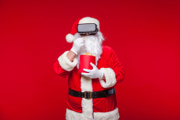 Santa Claus wearing virtual reality goggles and a red bucket with popcorn, on a red background. Christmas