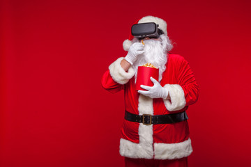 Santa Claus wearing virtual reality goggles and a red bucket with popcorn, on a red background. Christmas