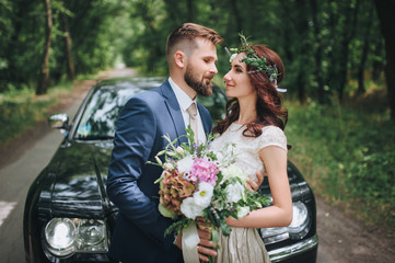 Newlyweds near the black wedding car stand on the road in the summer forest. A bride with a wreath...