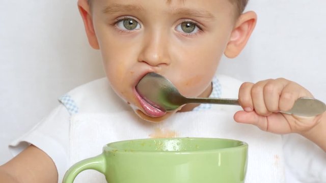 The child eats soup with a spoon from a plate close up. The boy puts the spoon in his mouth.