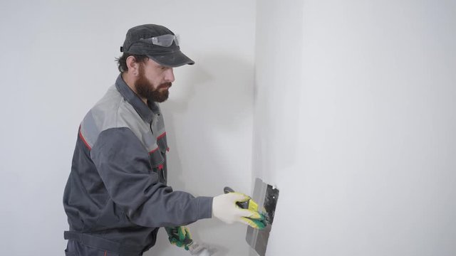 An adult builder with a beard aligns the wall with a long spatula and plaster, the man applies the composition to the surface