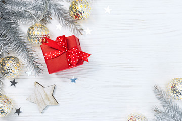 red gift box and silver christmas fir tree branches decorated with glowing garland on white wooden background with star confetti