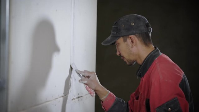 A young man who works as a builder puts a white grout on the joints between the tiles, he uses a spatula