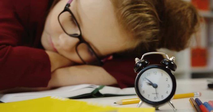 Close up of the cute schoolgirl sleeping on the textbook at the desk with school stuff. Alarm clock in the front. Inside