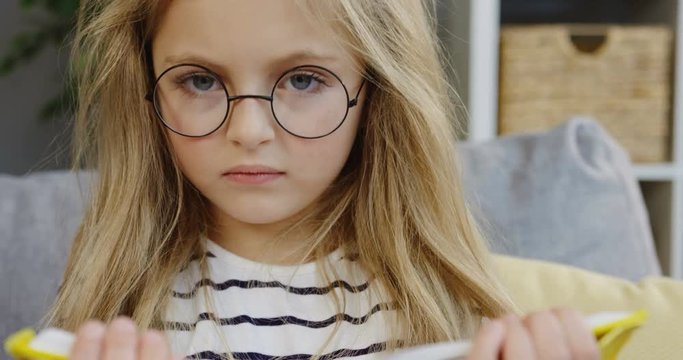 Close up of the funny blonde little girl in glasses closing a book and starting to grimase. Portrait. Indoor