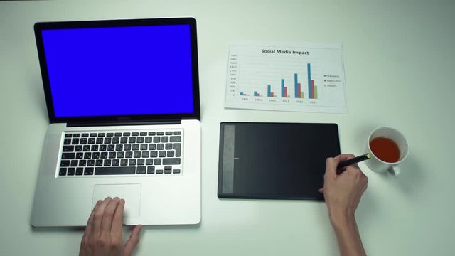 Top view male hands using graphic tab and laptop with green screen at white desk
