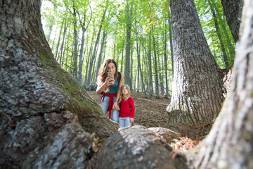 mother woman holding hand four years age blonde child with red shirt and blue jeans and taking a photo with mobile smartphone to a chestnut trunk in autumn forest
