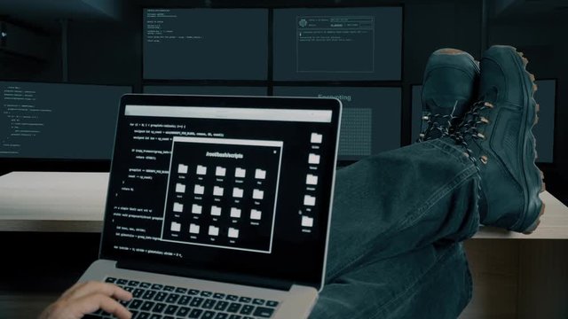 Hacker feet up to table. Hacker with laptop coding exploit to crack code. Tracing contact and satellite upling through internet. Man in military shoes sits and feet up on desk. Laptop computer screen.