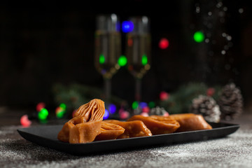 New Year's still-life. Glasses with champagne, New Year's lights, Christmas tree and sweet pastry. On a black plate