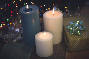 Obraz na płótnie Canvas A festive still life with a light blue and two white candles of different size and a handcrafted gift box with a turquoise bow on a colored wooden surface. A blurred Christmas tree with a garland and