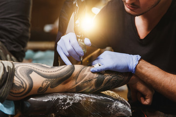 Close up of professional tattooer artist doing picture on hand of man by machine black ink from a jar. Tattoo art on body. Equipment for making tattoo art. Master makes tattooed in light studio. - obrazy, fototapety, plakaty