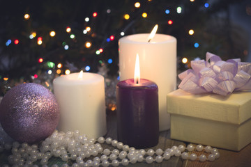 Obraz na płótnie Canvas A festive still life with a purple and two white candles of different size, a beige gift box with a lilac bow, white pearl beads and a lilac shiny ball. Dark Christmas background. Blurred bokeh