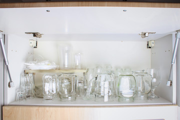 Different glassware in a white closet on the shelf. Banks, jugs, glasses, mugs.