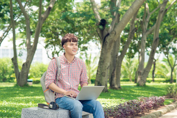 Naklejka na ściany i meble Attractive male asian student is using computer in university's park