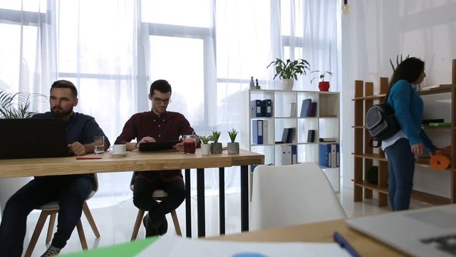 Beautiful asian young woman with yoga mat coming to work in open space freelance office. Sporty girl coming to workplace with exercise mat while her busy colleagues working with gadgets on background.
