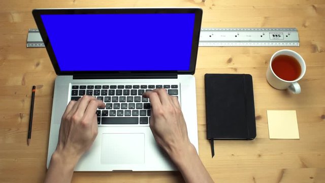 Young man using laptop with green screen at wooden desk. Top view on male hands
