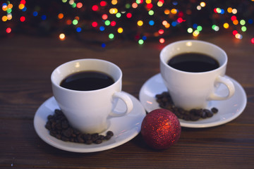 A festive still life with two white porcelian cups of hot black coffee. Coffee beans scattered on the saucers. A glitter red Christmas-tree ball in a close up. A rustic wooden background with