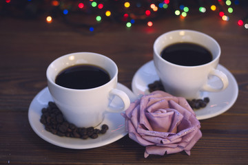 A festive still life with two white porcelian cups of hot black coffee. Coffee beans scattered on the saucers. A lilac paper rose in a close up. A rustic wooden background with Christmas lights. Dark