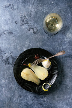 Traditional dessert sliced poached pears in white wine served in black plate with syrup, ice cream, lemon zest and glass of white wine over blue texture background. Top view with space