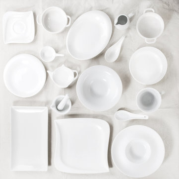 Big set of empty white porcelain plates and other tableware different size and shapes over white linen tablecloth. Flat lay