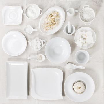 Big set of empty white porcelain plates and other tableware different size and shapes with white breakfast ingredients over white linen tablecloth. Flat lay