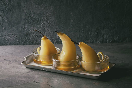 Traditional dessert poached pears in white wine served in glass bowls with syrup and lemon zest over gray texture table.