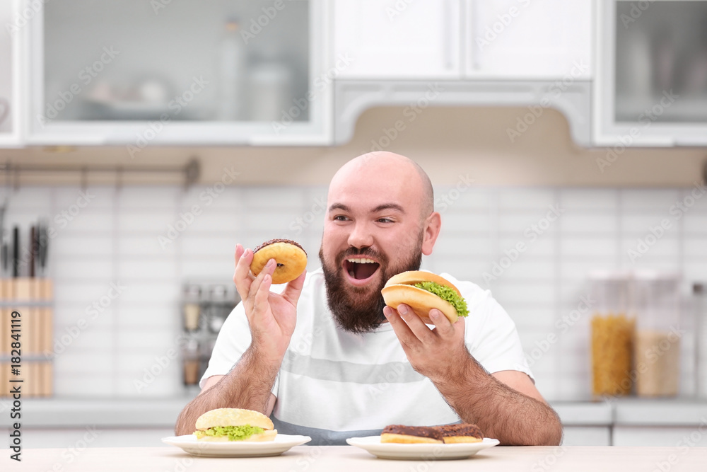 Wall mural Overweight young man with unhealthy food at table in kitchen