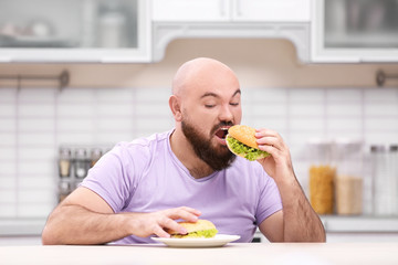 Overweight young man with unhealthy food at table in kitchen