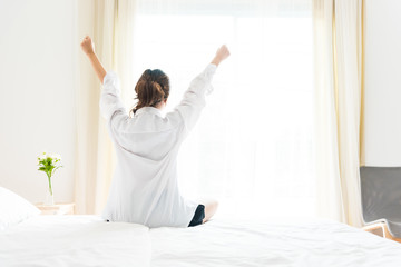 Back view of woman stretching in morning after waking up on bed near window. Holiday and Relax concept. Lazy day and Working day concept. Office woman and worker in daily life theme