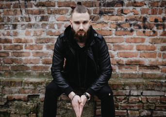 Modern young bearded man in black style clothes posing against brick wall.