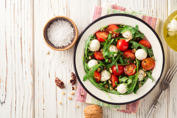 Fresh salad with arugula, cherry tomatoes, mozzarella cheese and walnuts on white wooden background.