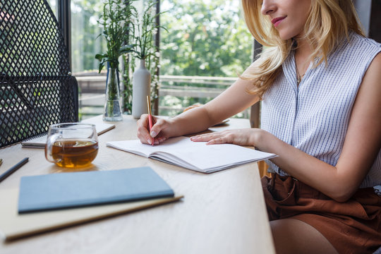 Cropped blonde Caucasian woman writing in her notebook at home office.