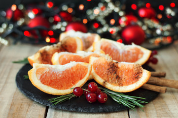 Citrus dessert. Orange fruit with cinnamon powder, green rosemary leaves and red currant on black slate board, Christmas lights and decor as background. Healthy food. Winter dessert