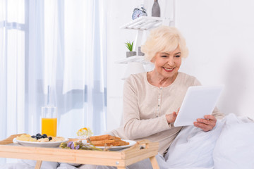 happy senior woman with breakfast in bed using tablet at home