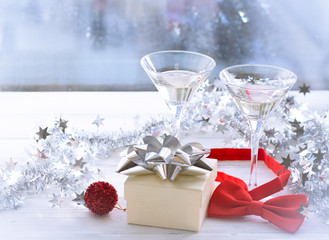 A romantic festive afternoon with two glasses of chapmagne, a burgundy gift box with a silver shiny bow. A red textile bow tie and a silver shiny garland on a white wooden table. A blurred window view
