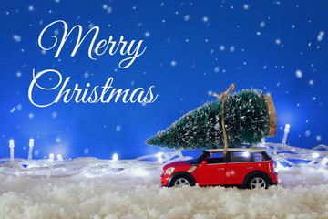 Red car carrying a christmas tree over the snow with garland lights.