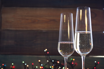 A festive still life with two glasses of champagne on a dark colored wooden background. Christmas fairy lights on the back. Close up. Dark colorful bokeh