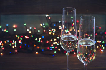 A festive still life with two glasses of champagne. Dark colored wooden background. Christmas fairy lights on the back. Close up. Dark colorful bokeh