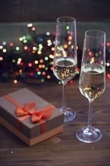 Two glasses of champagne and a brown gift box with an orange ribbon on a table.Dark colored wooden background. Christmas fairy lights on the back. Dark colorful bokeh