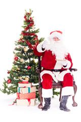 Portrait of Santa Claus sitting in chair next to Christmas tree, heap of gift boxes near him