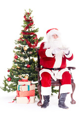 Portrait of Santa Claus sitting in chair next to Christmas tree, heap of gift boxes near him