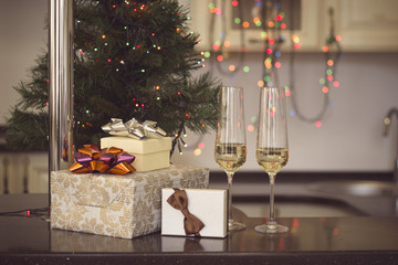Still life with two glasses of champaign and gift boxes with colorful bows on a black kitchen table. Christmas tree with colorful fairy lights in the back. Warm colors.